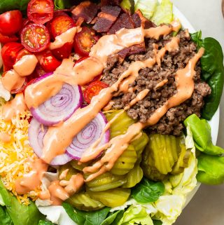 A cheeseburger in a bowl salad in a bowl with seasoned ground beef, grape tomatoes, red onions, pickles, turkey bacon and shredded cheese with special sauce