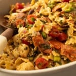 Mini bow tie pasta salad with pepperoni, salami, mozzarella pearls, cherry tomatoes, cucumber, olives, fresh herbs, and Italian dressing in a serving bowl.