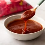 A small white bowl filled with smooth, vibrant red rhubarb barbecue sauce resting on a light marble surface.
