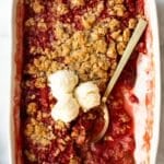 Overhead shot of the full baking dish of rhubarb crisp topped with three scoops of vanilla ice cream and a golden serving spoon.